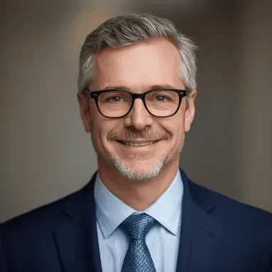 Tom B A smiling man in a suit and tie with glasses, standing against a neutral background.